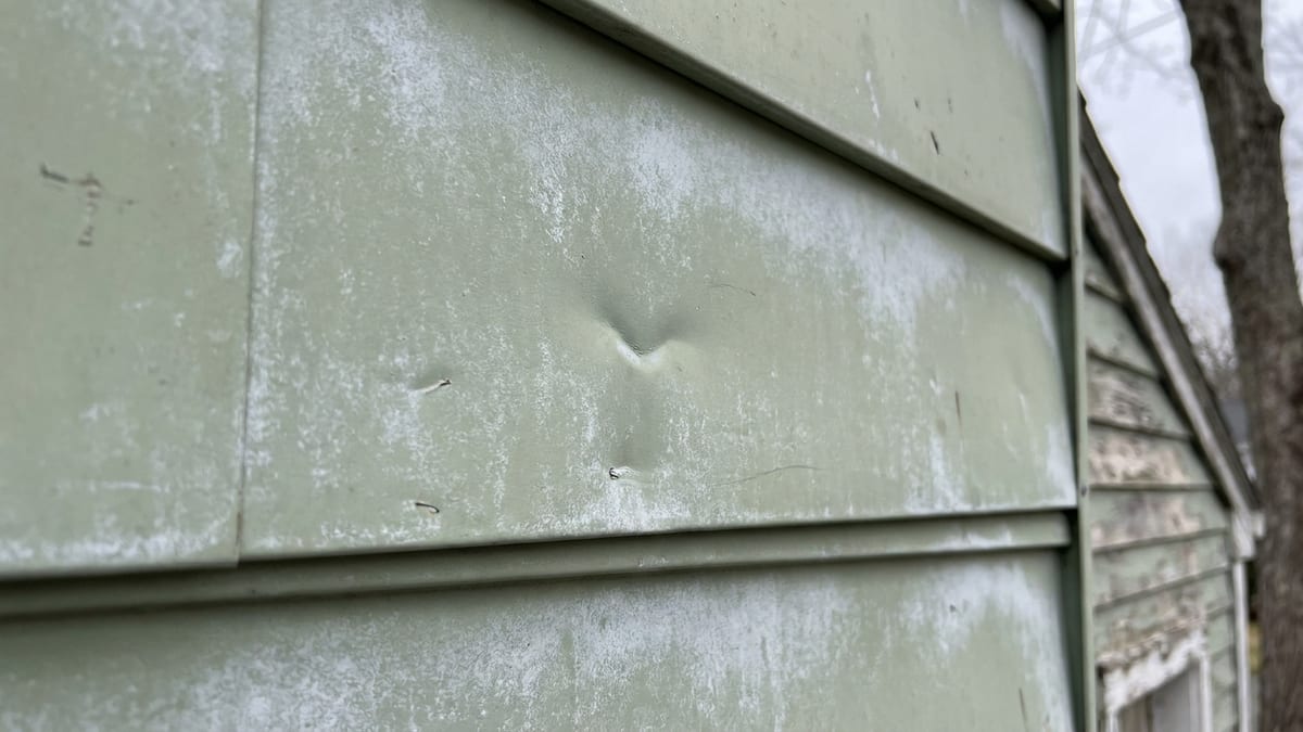 Close-up of chalky, oxidized pale aluminum siding on a mid-century ranch home with a small dent visible in flat daylight