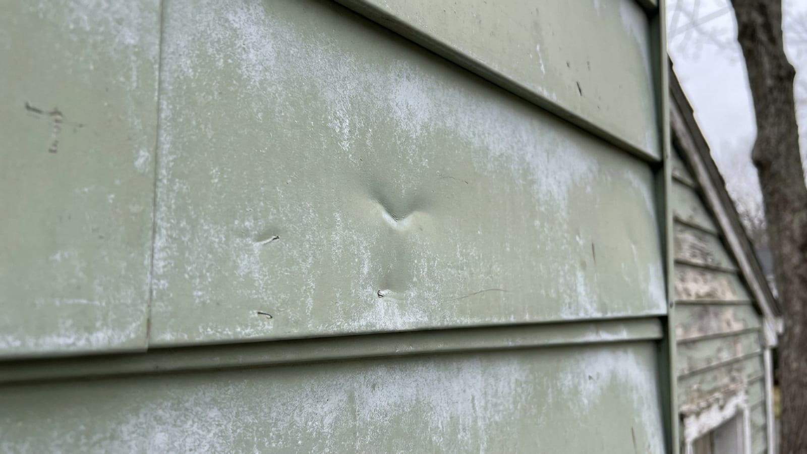 Close-up of chalky, oxidized pale aluminum siding on a mid-century ranch home with a small dent visible in flat daylight