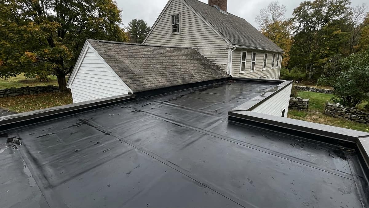 Low-slope black rubber membrane roof over a small home addition with visible seams and metal edge flashing, meeting an adjoining shingled roof and painted clapboard wall