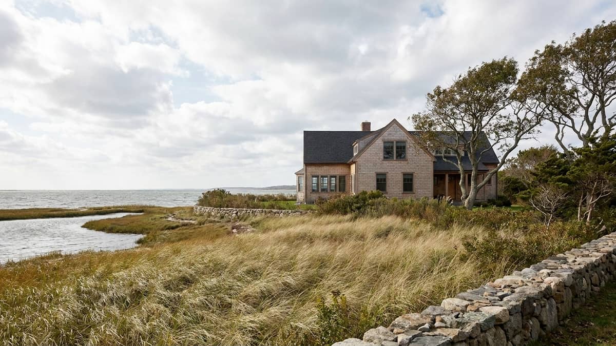 Shoreline Connecticut home with cedar-toned siding and an asphalt shingle roof, salt marsh grasses and open water behind it under a wide sky