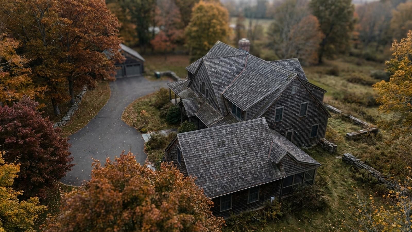 Aerial-angle view of a two-story suburban Connecticut house with a gabled and dormered roof in weathered gray shingles, autumn trees along the driveway