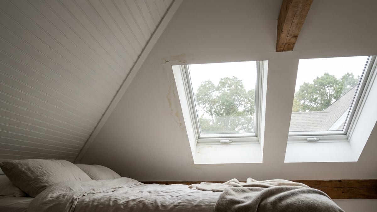 Interior of a bright vaulted attic bedroom looking up at two skylights set into a sloped white ceiling, with faint water staining on the drywall around one frame