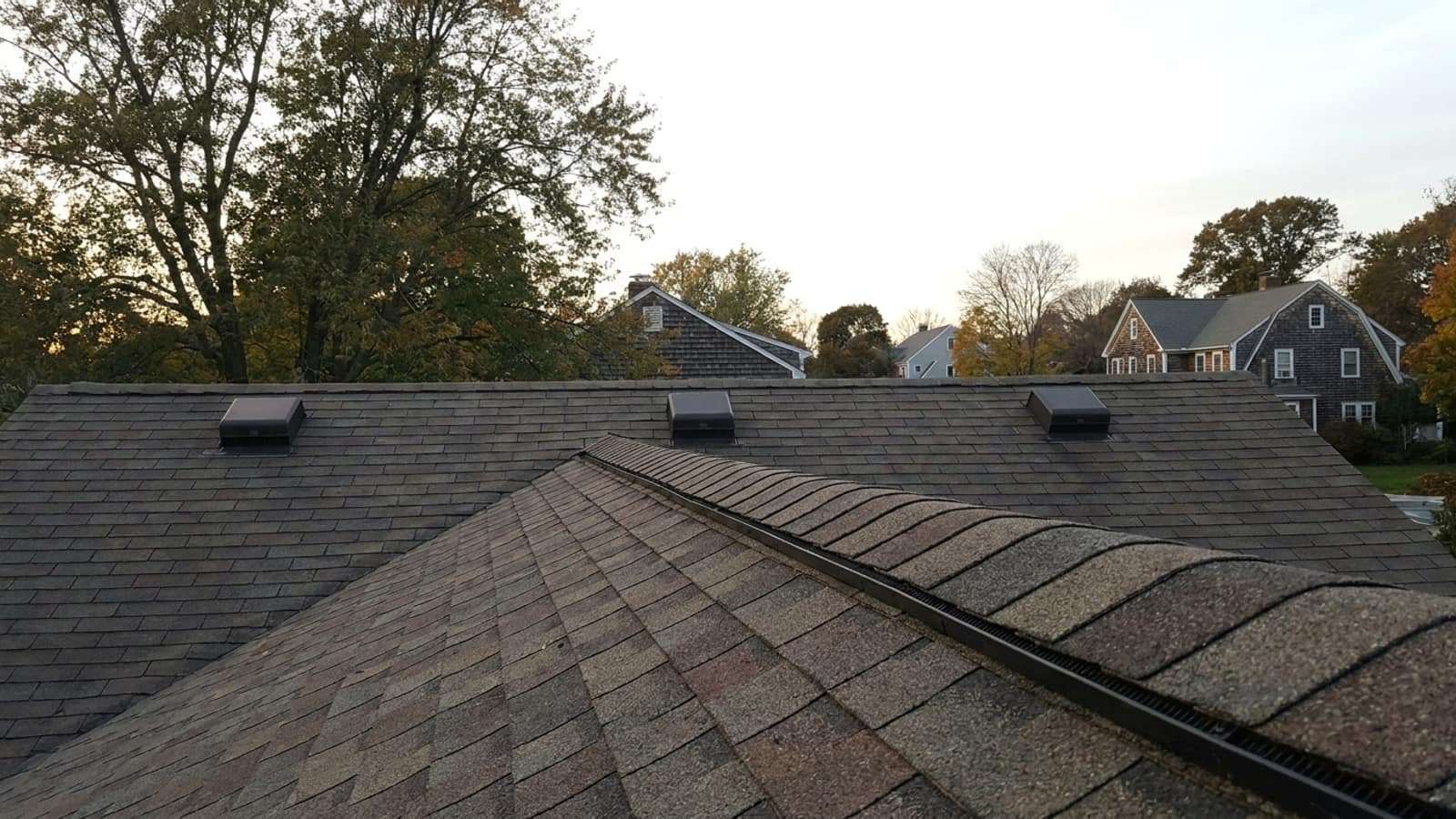 Close view of an asphalt shingle roof peak with a continuous shingle-over ridge vent, and square box vents on a second roof plane behind it in late afternoon light