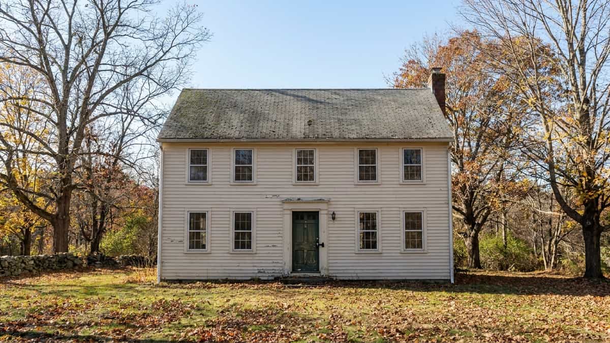 A two-story gray colonial house in Connecticut on a clear fall afternoon with aging asphalt shingles and a real estate lockbox on the front door