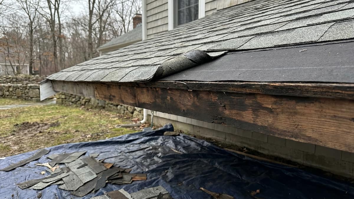 Close-up of a residential roof edge during replacement with weathered gray asphalt shingles peeled back to reveal wood decking and new underlayment