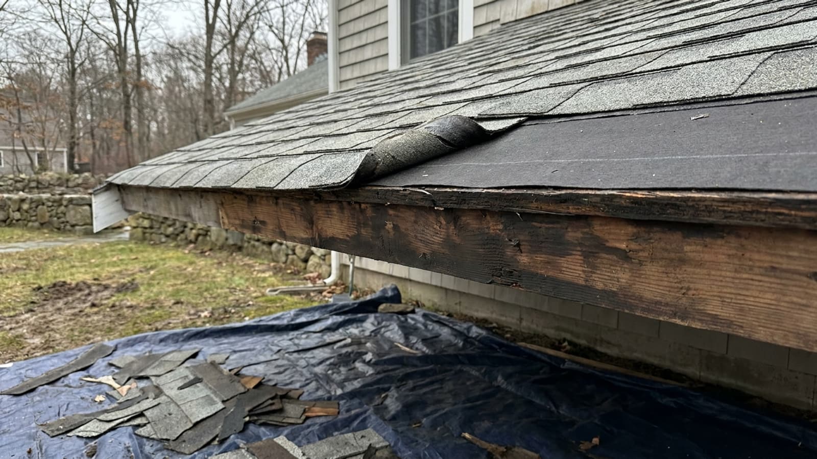 Close-up of a residential roof edge during replacement with weathered gray asphalt shingles peeled back to reveal wood decking and new underlayment