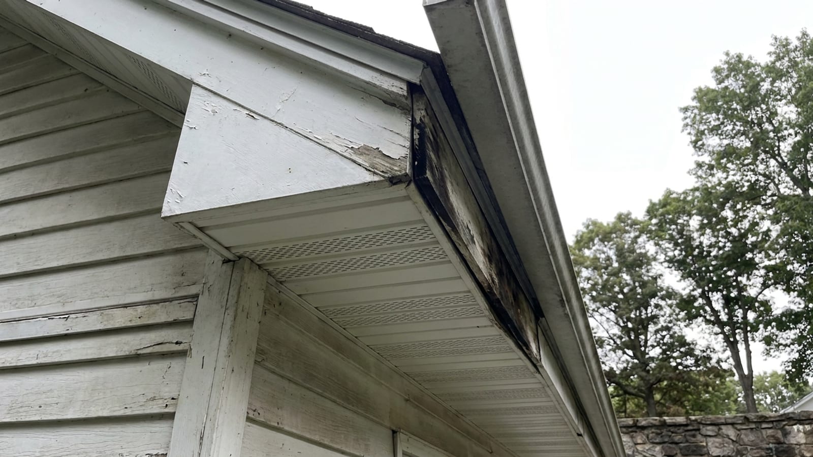 Upward view of a house eave with white vented aluminum soffit panels and a fascia board meeting the gutter, one section of fascia water-stained and separating, clapboard siding below and open sky past the roof edge