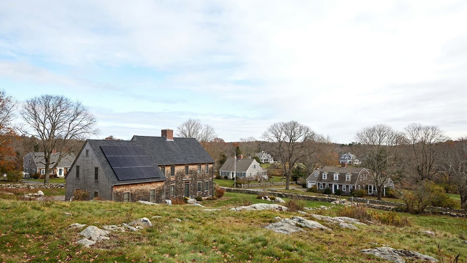 A suburban house seen from a neighboring hillside with a black solar array covering half of a south-facing asphalt shingle roof under a clear blue sky