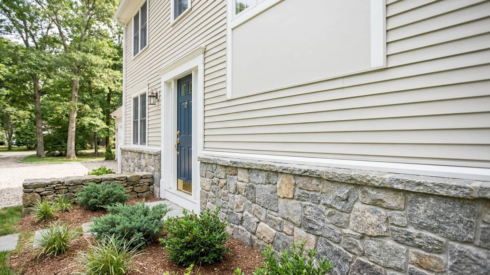 Stone veneer water table wrapping the base of a home beneath light vinyl siding beside a front entry in bright daylight