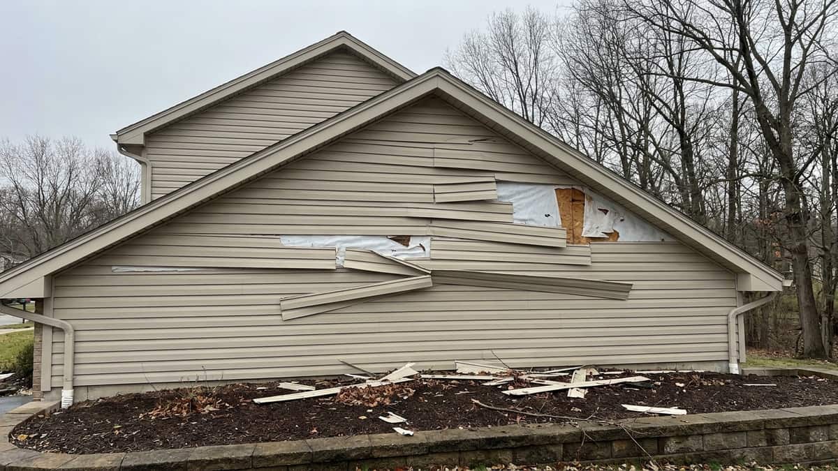 One gable wall of a suburban home with several vinyl siding panels cracked and pulled loose by wind, a few panels missing to expose house wrap, debris scattered on the mulch bed below under post-storm daylight