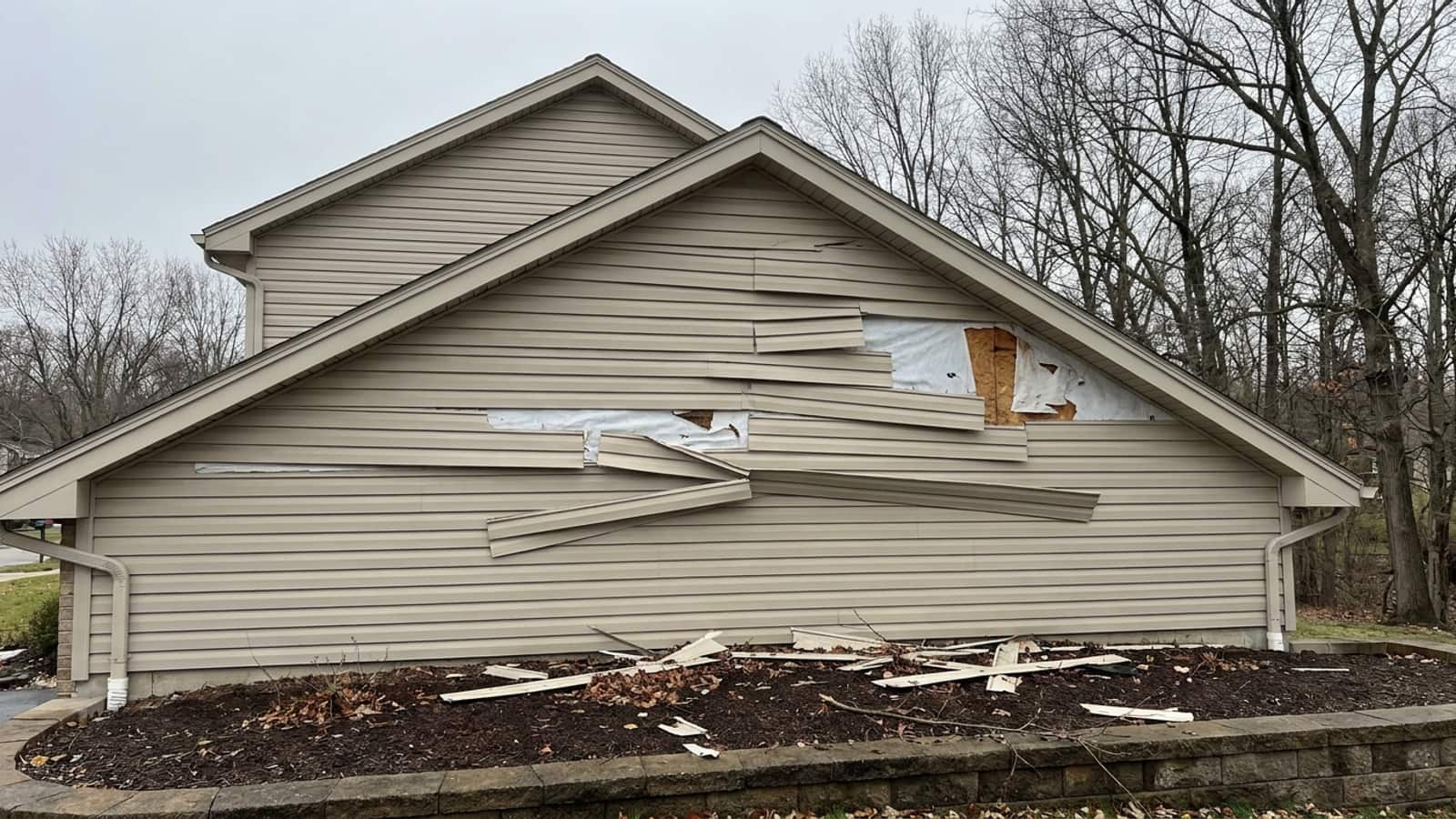 One gable wall of a suburban home with several vinyl siding panels cracked and pulled loose by wind, a few panels missing to expose house wrap, debris scattered on the mulch bed below under post-storm daylight