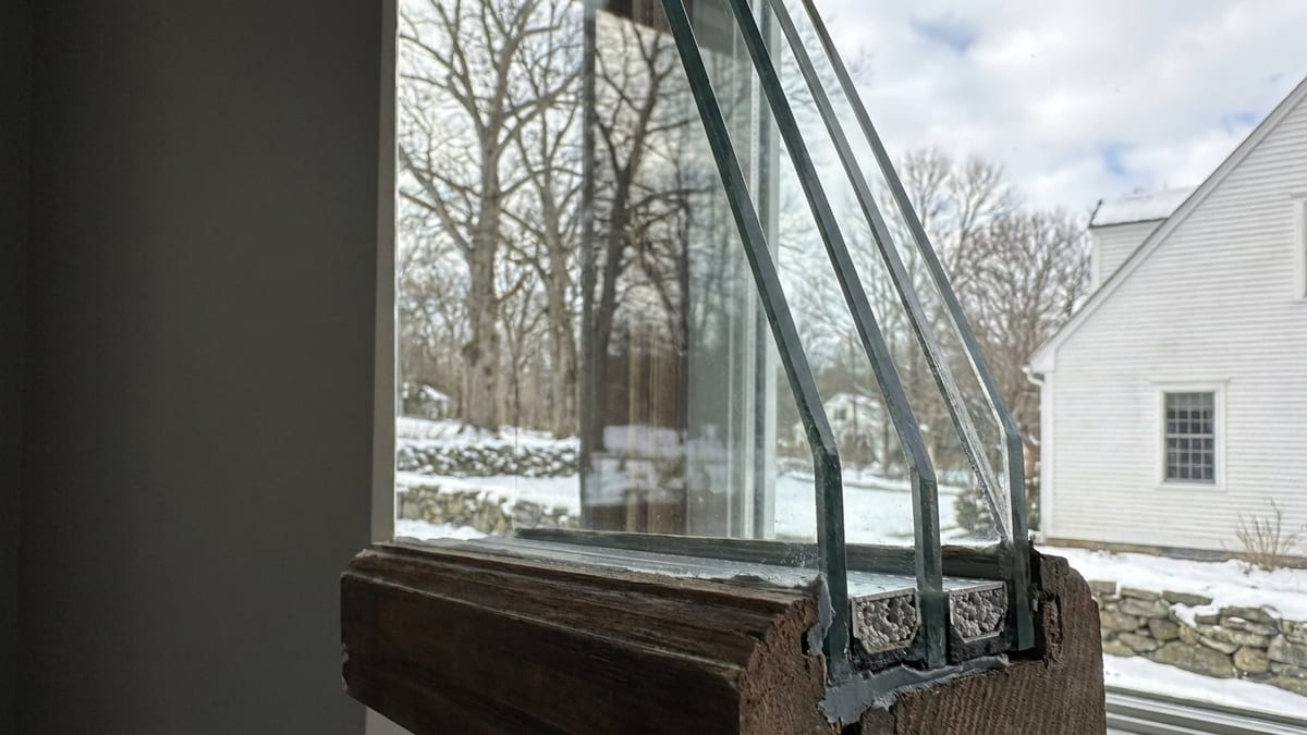 Cutaway edge of a triple-pane window sash showing three glass layers, two sealed air spaces and the spacer bars between them, with a winter yard reflected in the glass