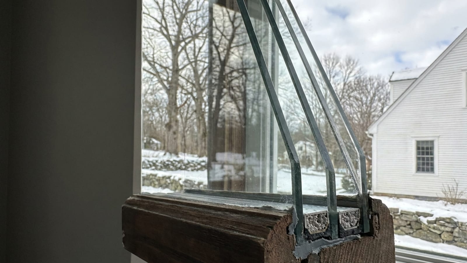 Cutaway edge of a triple-pane window sash showing three glass layers, two sealed air spaces and the spacer bars between them, with a winter yard reflected in the glass