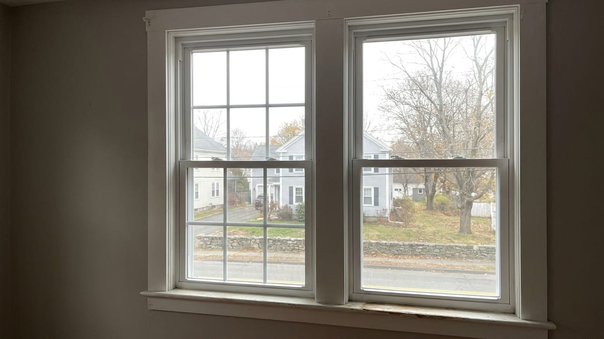 Two side-by-side white double-hung replacement windows in a neutral painted wall, one with colonial six-over-six grille bars and one with clear glass, lit by soft daylight