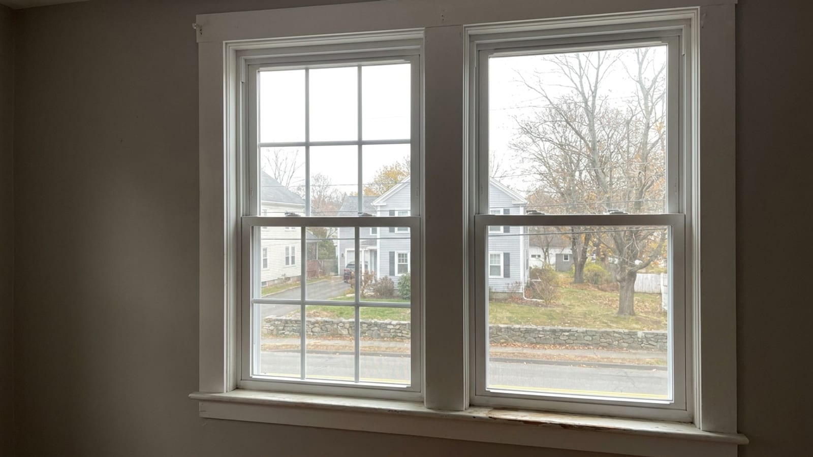 Two side-by-side white double-hung replacement windows in a neutral painted wall, one with colonial six-over-six grille bars and one with clear glass, lit by soft daylight