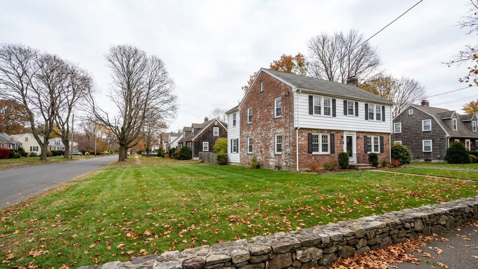 A brick and white clapboard 1930s colonial home with rows of white double-hung windows and autumn leaves on the front lawn