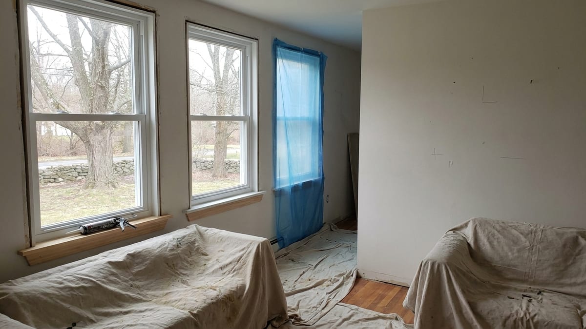 Living room with two newly installed white vinyl double-hung windows and a third opening covered in protective plastic sheeting, drop cloths on the hardwood floor and a caulk gun resting on the sill
